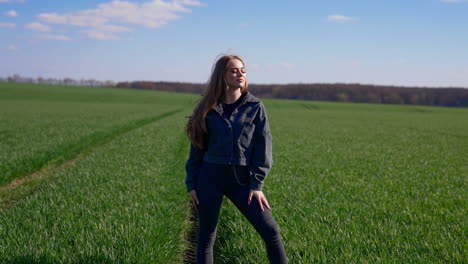 attractive girl posing on camera outdoors. beautiful young model in tight pants and cotton jacket with fluttering hair looking on camera on green field background.
