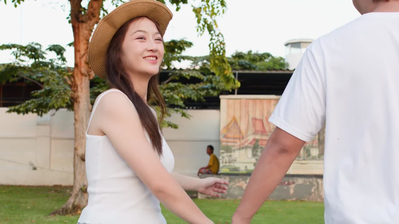 A happy Asian woman in a straw hat walks hand-in-hand with her boyfriend outdoors
