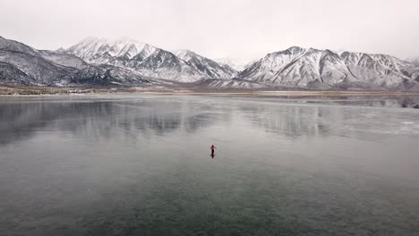 Eisfischen-Auf-Der-Zugefrorenen-Seeoberfläche-Am-Crowley-Lake-Im-Kalten-Winter-Kalifornien,-Person-Auf-Dem-Eisigen-See,-Die-Nach-Einem-Ort-Sucht,-Um-Ein-Loch-Zum-Eisfischen-Zu-Graben,-Landschaft-Am-See-Und-Schneebedeckte-Felsige-Berge-Am-Horizont