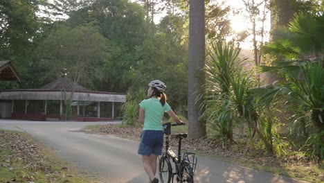 A dynamic panning shot of a female cyclist walking her bicycle along the street while looking around the houses of the neighborhood. She's walking towards the junction and the sun is starting to set