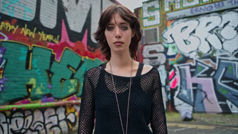 outdoor fashion portrait of young alternative style woman leaning in smiling into camera against graffiti covered walls of london city street uk in real time