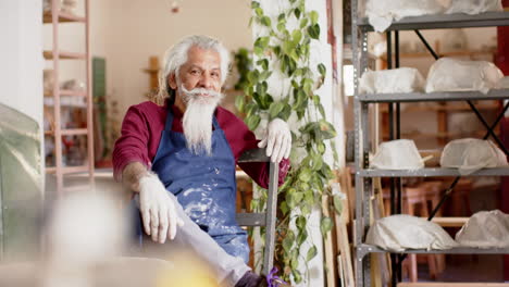 happy senior biracial potter with long beard resting and smiling in pottery studio, slow motion