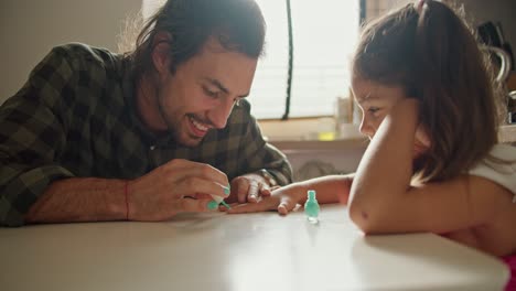 A-brunette-man-in-a-green-checkered-shirt-gives-his-little-daughter-a-brunette-girl-in-a-pink-dress-a-manicure-using-green-polish-in-the-kitchen-on-the-table