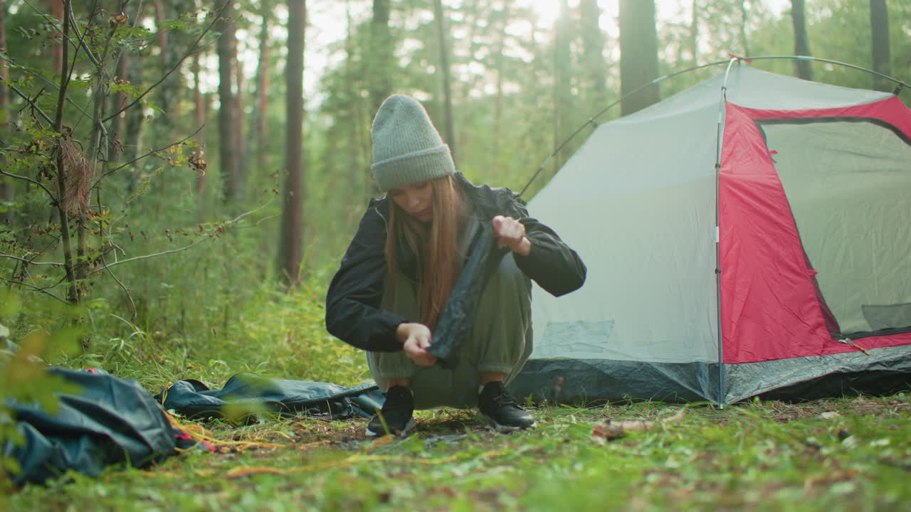 woman in gray beanie squats beside camping tent picking scattered tent rods from forest ground as morning sunlight gently filters through surrounding trees during peaceful outdoor activity