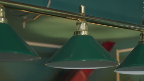 toma de primer plano de dos lámparas aéreas con sombras verdes en una sala de billar, destacando sus accesorios metálicos dorados y diseño elegante, arrojando luz suave en la mesa de billar debajo