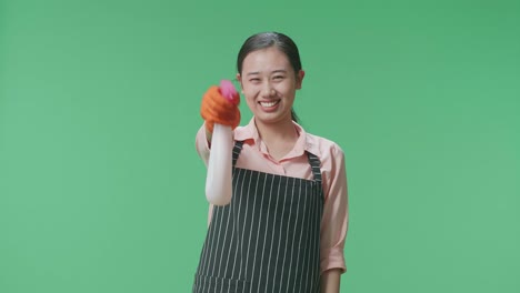 una ama de casa asiática sonriente con un delantal usando un spray mientras está de pie en el estudio de pantalla verde