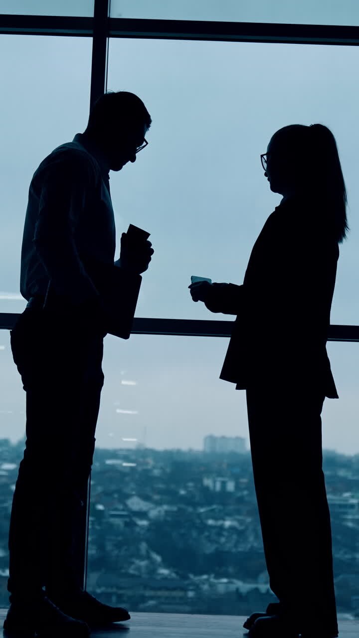 Silhouette scure di uomini e donne in piedi davanti alla finestra panoramica. Conversazione tra colleghi in ufficio. Angolazione dal basso. Video verticale