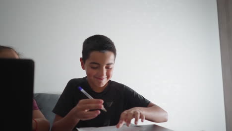 boy smiling proudly shows his written work to friend seated beside him in bright room while continuing to write with enthusiasm and joy, sharing fun moment of learning