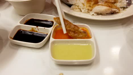 Chopsticks dip a piece of Hainanese chicken into vibrant sauces beside rice on a well-lit table, captured in a smooth, close-up sequence