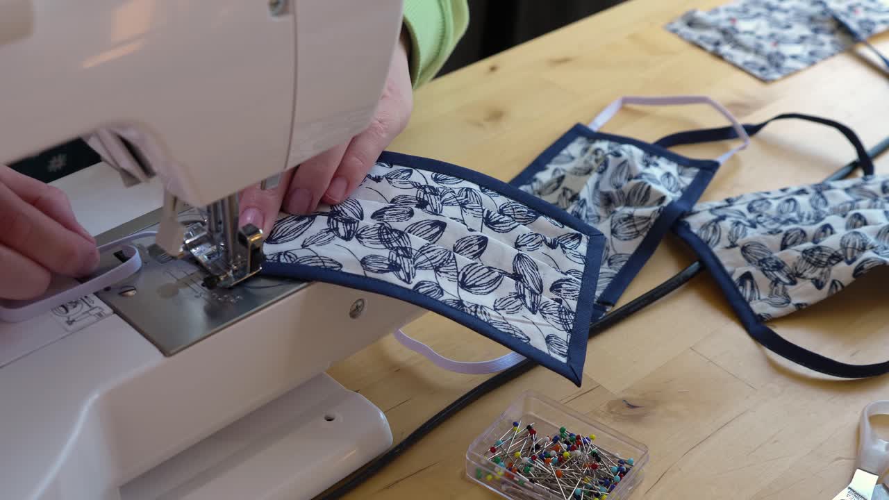 Woman sewing rubber band on a face mask with sewing utensils and finished masks in the background.
