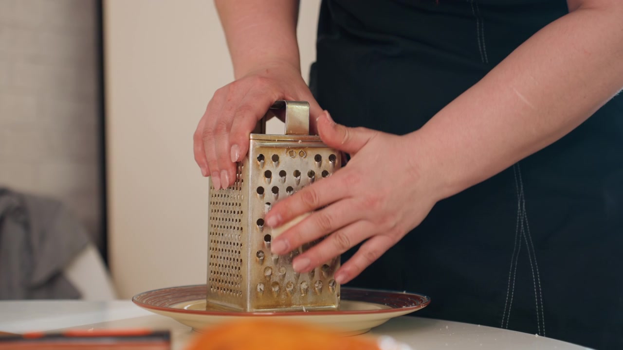 Close view of hands grating orange carrot against stainless steel box grater above shallow bowl on kitchen countertop, showing fresh food prep, texture, and steady cooking action, with hand, grating