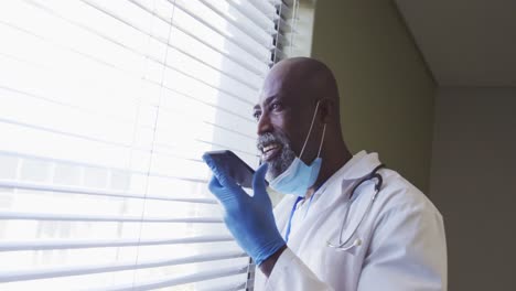 Portrait-of-african-american-male-doctor-wearing-face-mask-talking-using-smartphone