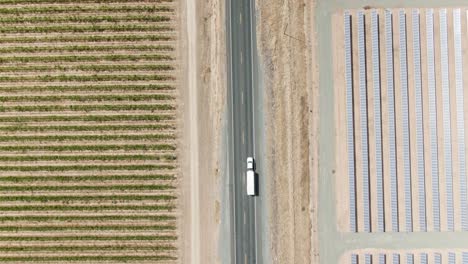 Vista-De-Pájaro-A-Gran-Altitud-Sobre-Una-Carretera-Rural-Local-Rodeada-De-Paneles-Solares-En-El-Lado-Derecho-Y-Un-Viñedo-En-El-Lado-Izquierdo