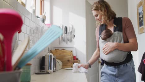 Video-of-caucasian-mother-with-newborn-baby-in-baby-carrier-cleaning-kitchen