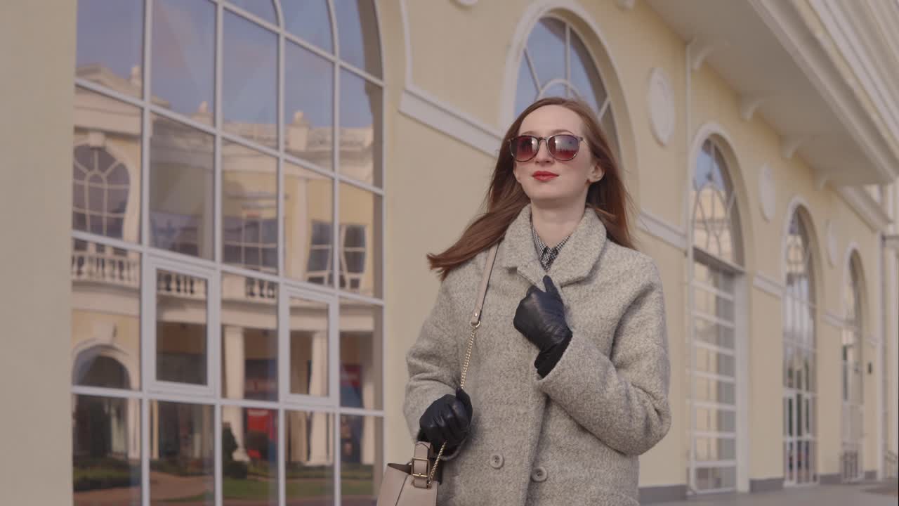 베이지 코트 를 입은 여자 가 현대적 인 건물 밖 에서 고 있다