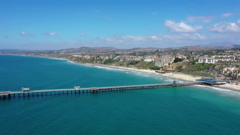 aerial 4k slider shot 45-degree towards san clemente pier in orange county, california while people eat at restaurants, sunbathe on beach, fishing, and surfing