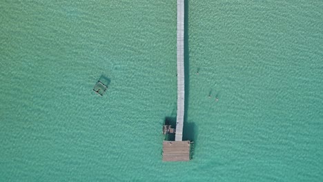 Aerial,-top-down,-birdseye,-drone-shot-of-a-wooden-pier-and-people-swimming-in-the-turquoise-sea,-at-a-paradise-beach,-on-a-sunny-day,-in-Koh-Kood,-Thailand,-Asia