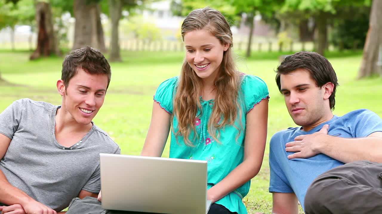 En kvinne ler og snakker mens hun bruker en laptop, sittende med sine to venner i en park.