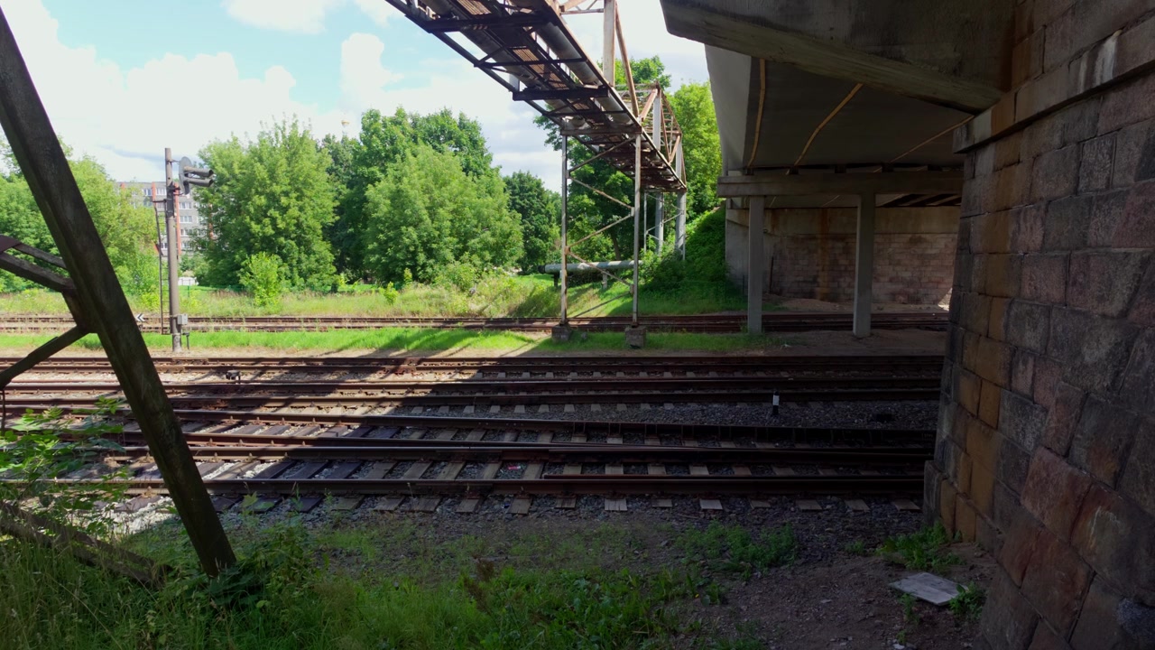 Wide shot showing multiple railway tracks passing under concrete bridge and metal pipe structure surrounded by green trees in Daugavpils Latvia during sunny summer.