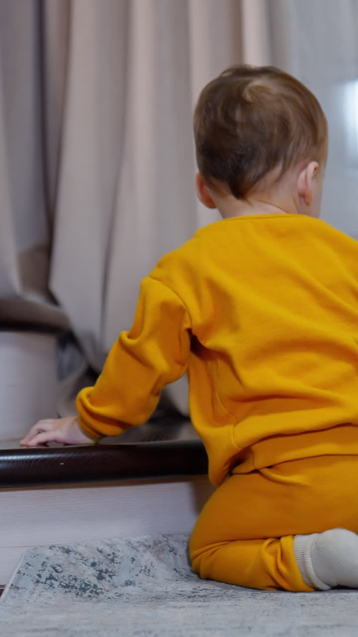 Lovely toddler in orange sport suit sits on the floor near the stairs. Kid throws the toy and watches it roll. Vertical video