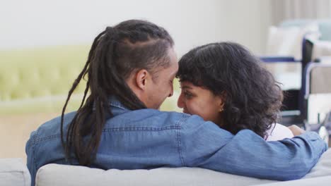 feliz pareja biracial sentada en el sofá riendo y abrazándose en la sala de estar