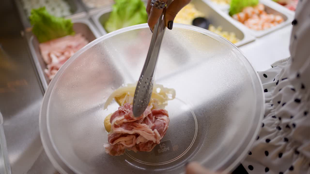 Person uses tongs to pick raw meat and vegetables at a well-lit hotpot buffet station