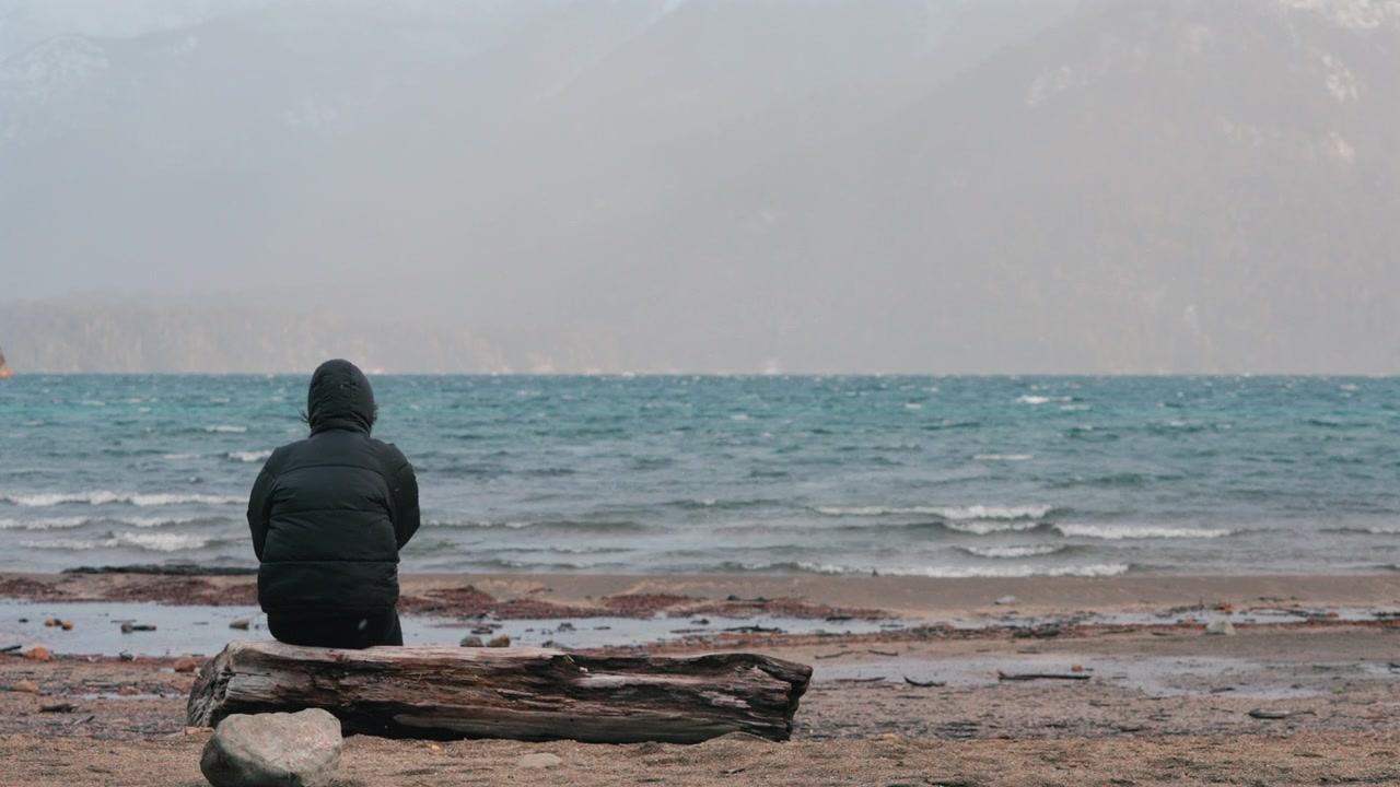 Una persona in cappotto invernale siede su un tronco, di fronte al mare agitato sotto cieli coperti. Ideale per temi di solitudine, contemplazione o viaggi invernali.