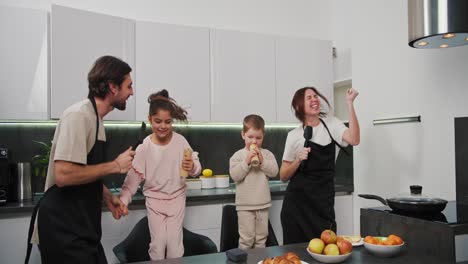 happy family brunette man with stubble in a black apron together with his brunette wife and two small children dancing and having fun in a modern kitchen before starting breakfast in a modern apartment in the morning
