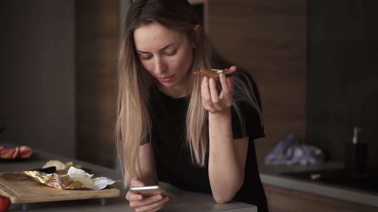 retrato de una mujer comiendo pan con mantequilla para el desayuno, revisando su teléfono inteligente en la cocina