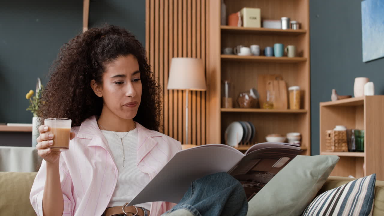 A woman relaxes on a couch, reading a magazine and holding a drink.