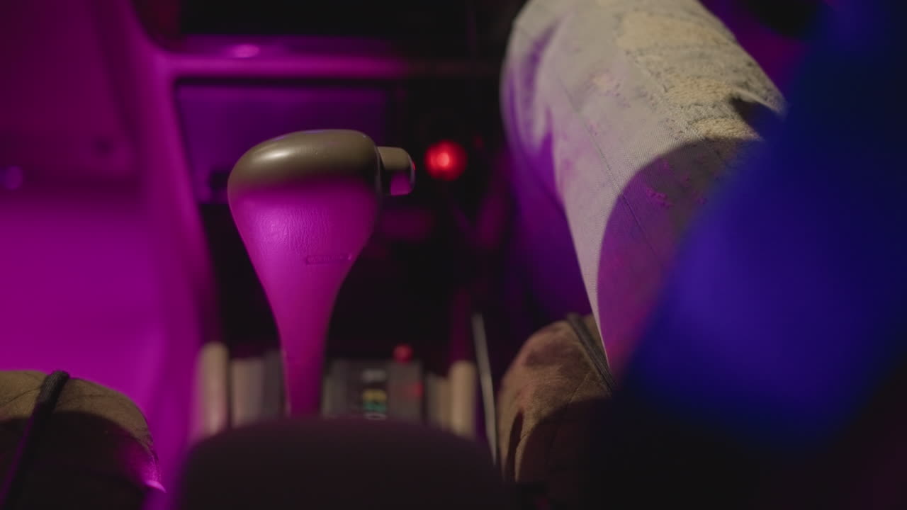 abitacolo notturno dell'auto, mano che stringe la console vicino alla leva del cambio, bagliore magenta neon del cruscotto, GPS di navigazione e percorso riflessi sullo schermo, pacco sul sedile del passeggero, concentrazione determinata del corriere