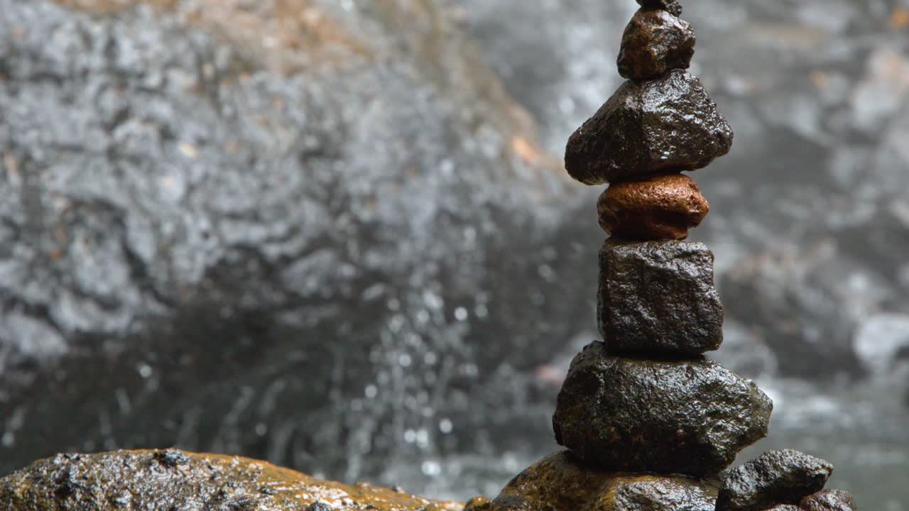 Inquadratura ravvicinata di rocce in equilibrio su massi fluviali bagnati, con uno sfondo di cascata sfocato.
