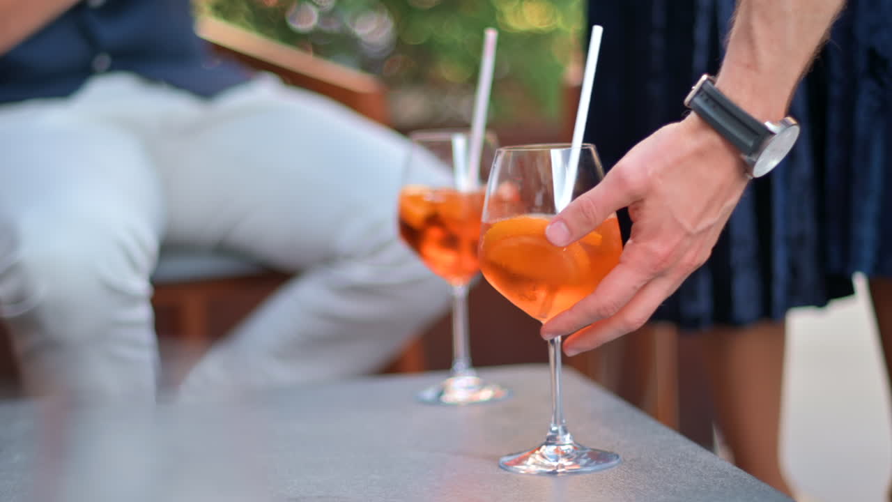 Arka planda insanların hareket ettiği bir terasta, masanın üzerinde iki kadeh aperol spritz