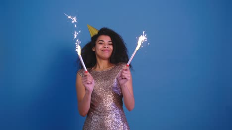 hermosa joven africana rizada en gorra de cumpleaños sosteniendo dos fuegos artificiales y divertirse