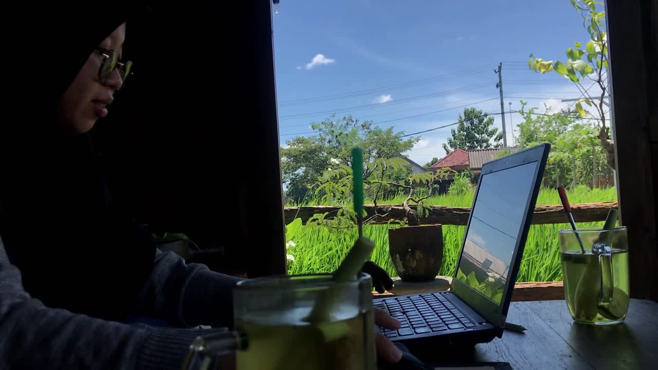 Muslim Woman Working on Laptop in Traditional Javanese Cafe with Green Rice Field View south east asian young woman, traditional wooden house, nature background
