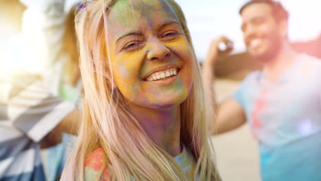 close-up portrait of a beautiful blonde girl dances in celebration of holi festival with her friends. her face and clothes are covered with colorful powder.