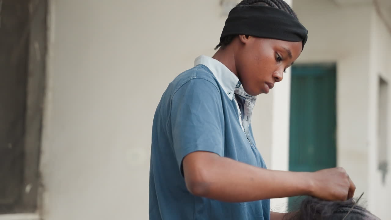 Young woman focuses on hair styling. Apprentice diligently braids hair outside in courtyard. Young stylist carefully works on tight hair sections outdoors in courtyard during community event