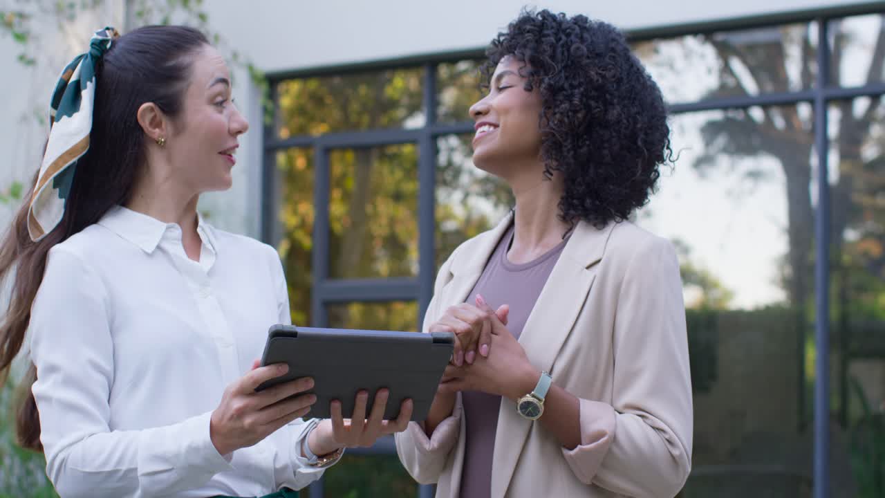Diverse female coworkers in work attire reviewing tablet report for meeting by glass doors smiling