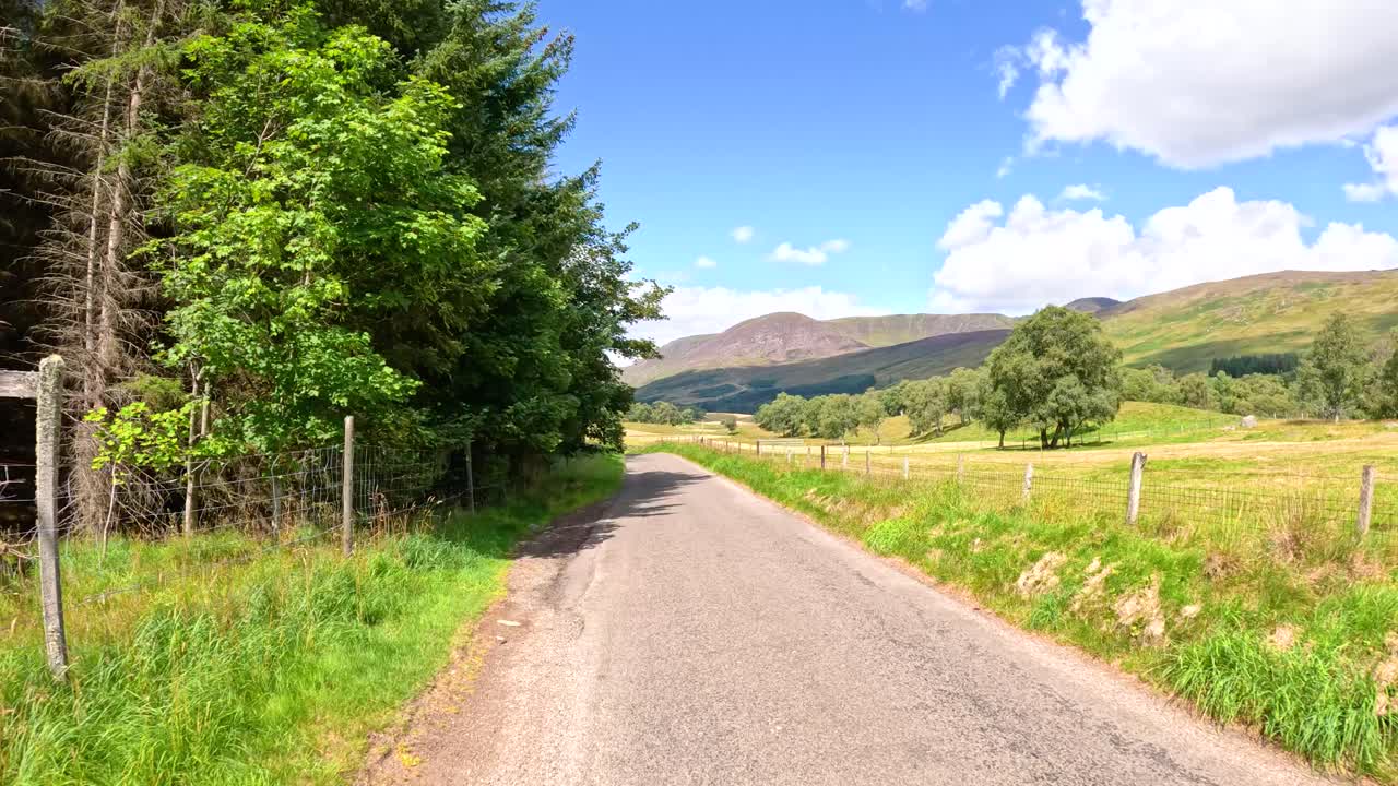Car drives along narrow rural road through green hills, trees, and farmland in Clova, Scotland