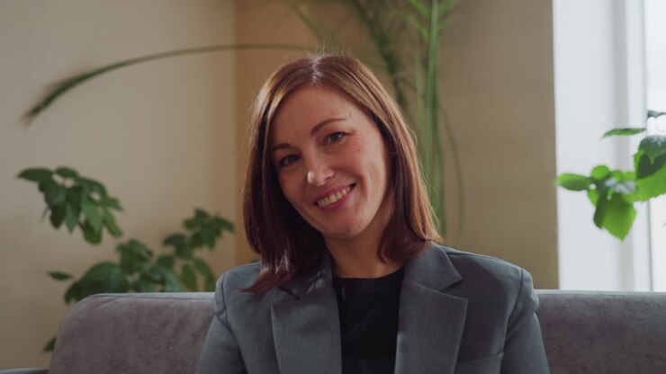 Confident professional woman in formal blazer sitting on sofa smiling warmly during interview or casual conversation in bright modern office surrounded by green plants and natural daylight