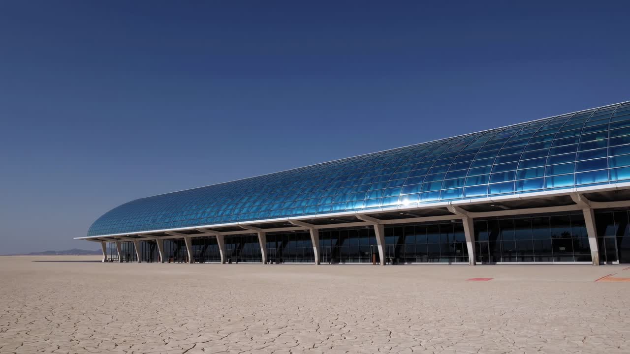 Modern building with a curved blue glass roof stands in a dry, cracked desert landscape under a clear blue sky, showcasing the contrast between nature and architecture