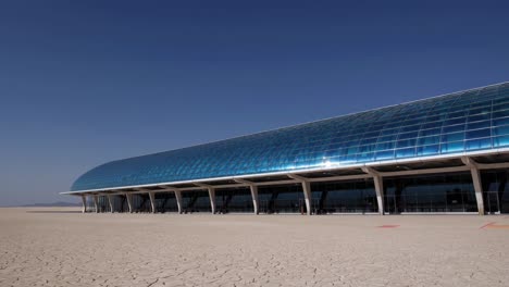 modern building with a curved blue glass roof stands in a dry, cracked desert landscape under a clear blue sky, showcasing the contrast between nature and architecture