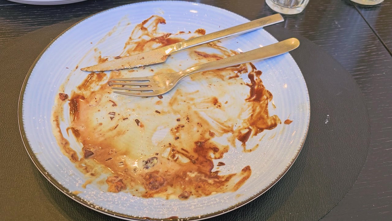 Empty dessert plate after eating a chocolate waffle, with fork and knife, melted chocolate sauce, cream smears, powdered sugar, crumbs, and cafe table in a casual restaurant setting indoors.
