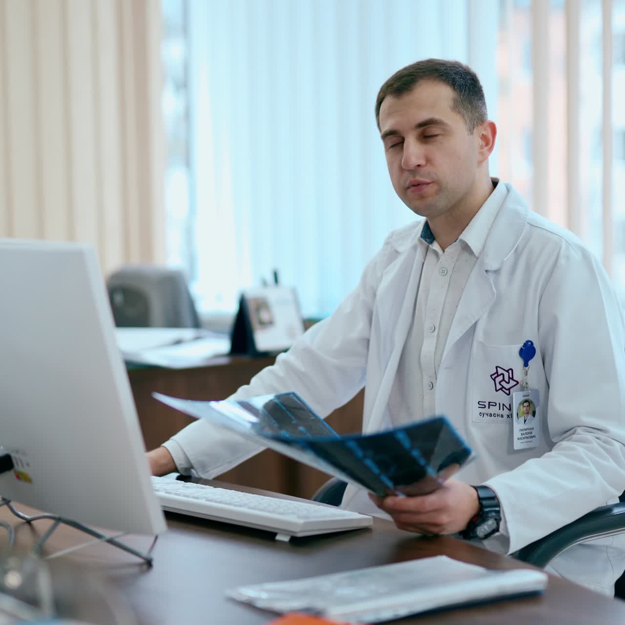 Portrait of a man doctor sitting in his cabinet. Specialist in white robe sits at the table in medical office and holds x-ray photograph.