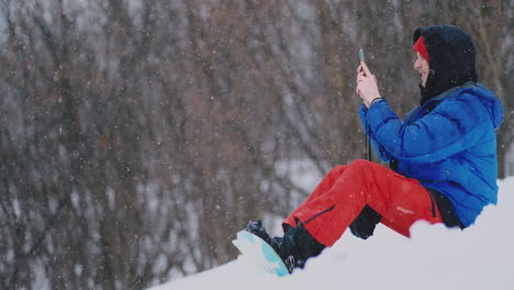 Male-snowboarder-sitting-on-the-snow-taking-photos-on-the-phone-beautiful-scenery-of-the-resort-for-social-networks