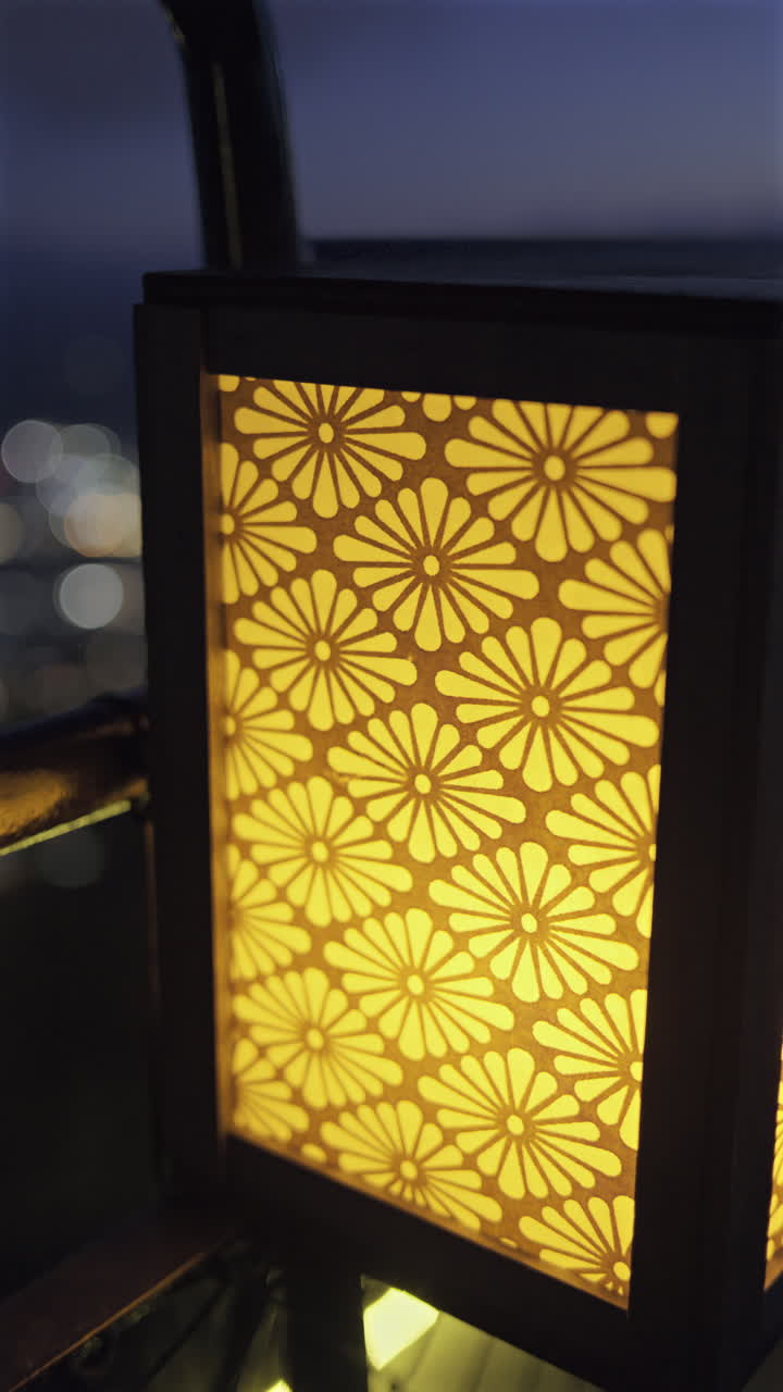 A beautiful floral lantern emits a warm glow on a balcony, creating a cozy atmosphere as city lights twinkle in the distance during the evening hours. Vertical Video