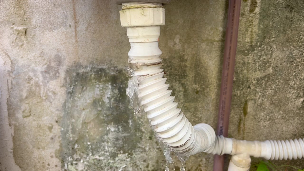 Close-up of a damaged white pipe leaking a stream of water against a weathered concrete exterior wall.