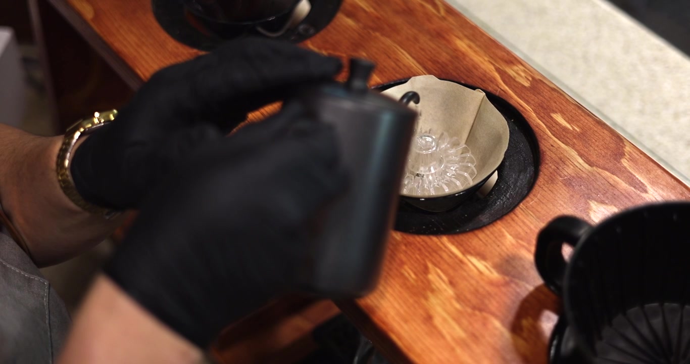 Mani che preparano caffè pour-over con filtro di carta e bollitore su un bancone di legno, mescolando lentamente per inumidire il fondo