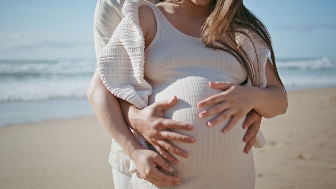 임신 한 가족이 해변에서 휴식을 취하고 배를 안고 가까이에서 남편이 아내를 포옹하고 있습니다.