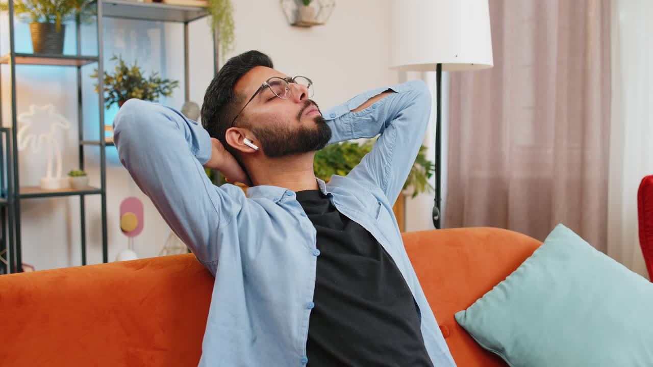 Happy relaxed indian man resting on home couch with wireless earphones listening ambient music